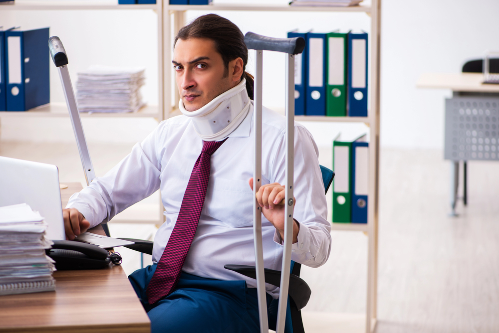 middle aged worker in neck brace with broken arm sitting at table and talking to businessman in blue jacket in office, compensation concept